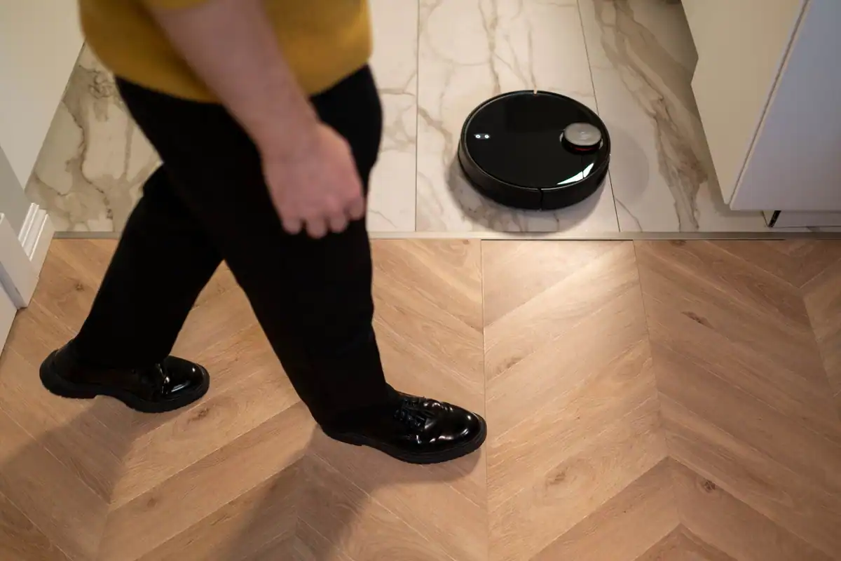Aspirator robot negru care curăță parchetul, în timp ce o persoană trece pe lângă el