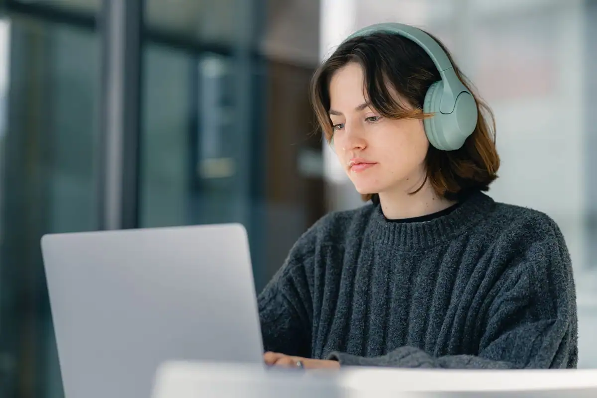 Tânără cu căști pe urechi lucrează concentrată la laptop, în interior