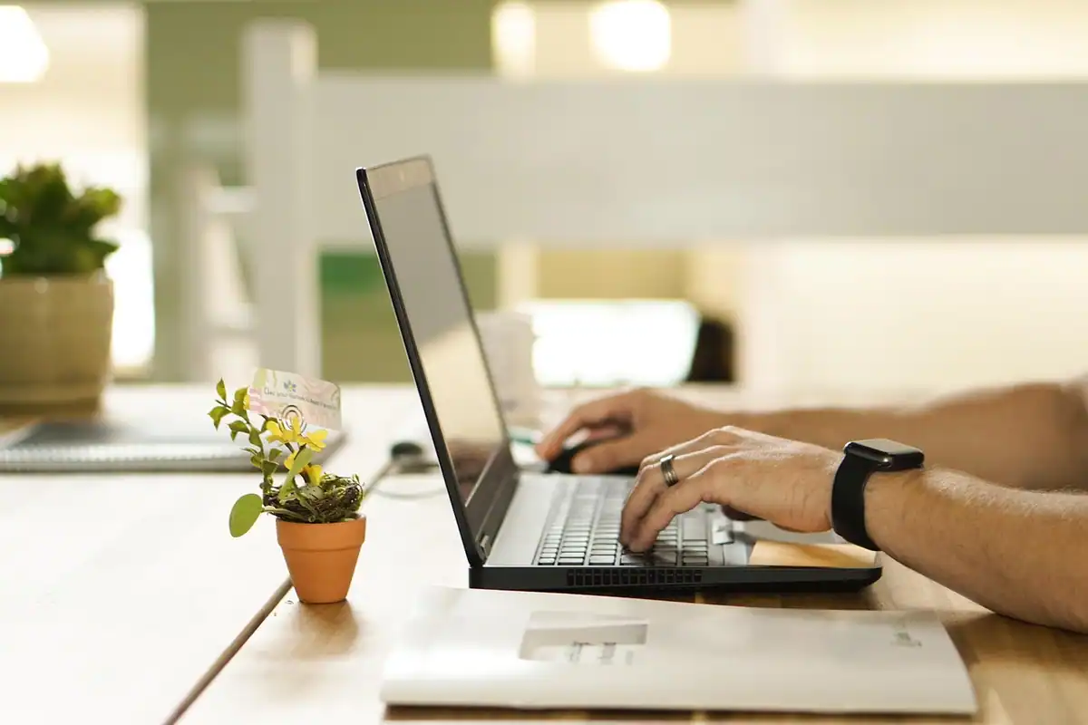 Bărbat care lucrează la laptop, cu un smartwatch negru la mână, lângă o plantă mică