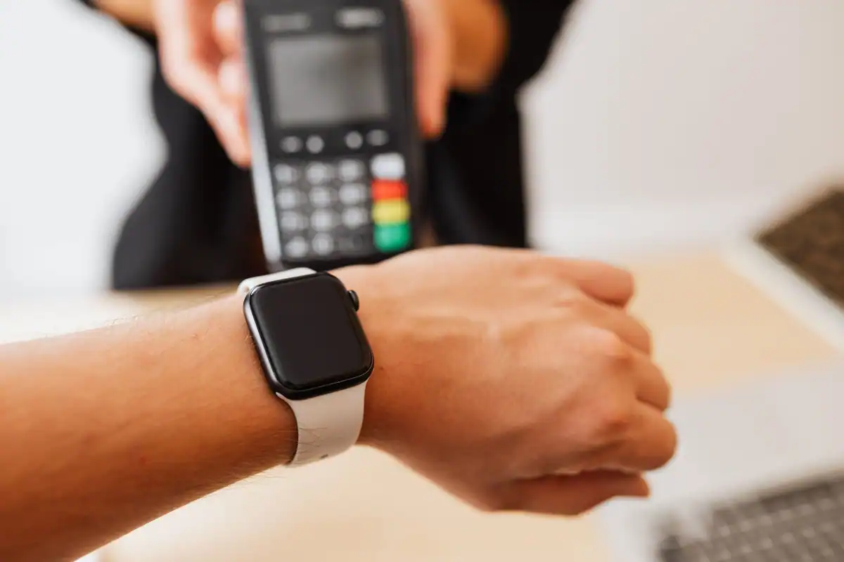 Smartwatch alb purtat la mână, în fața unui terminal de plată ținut de altă persoană