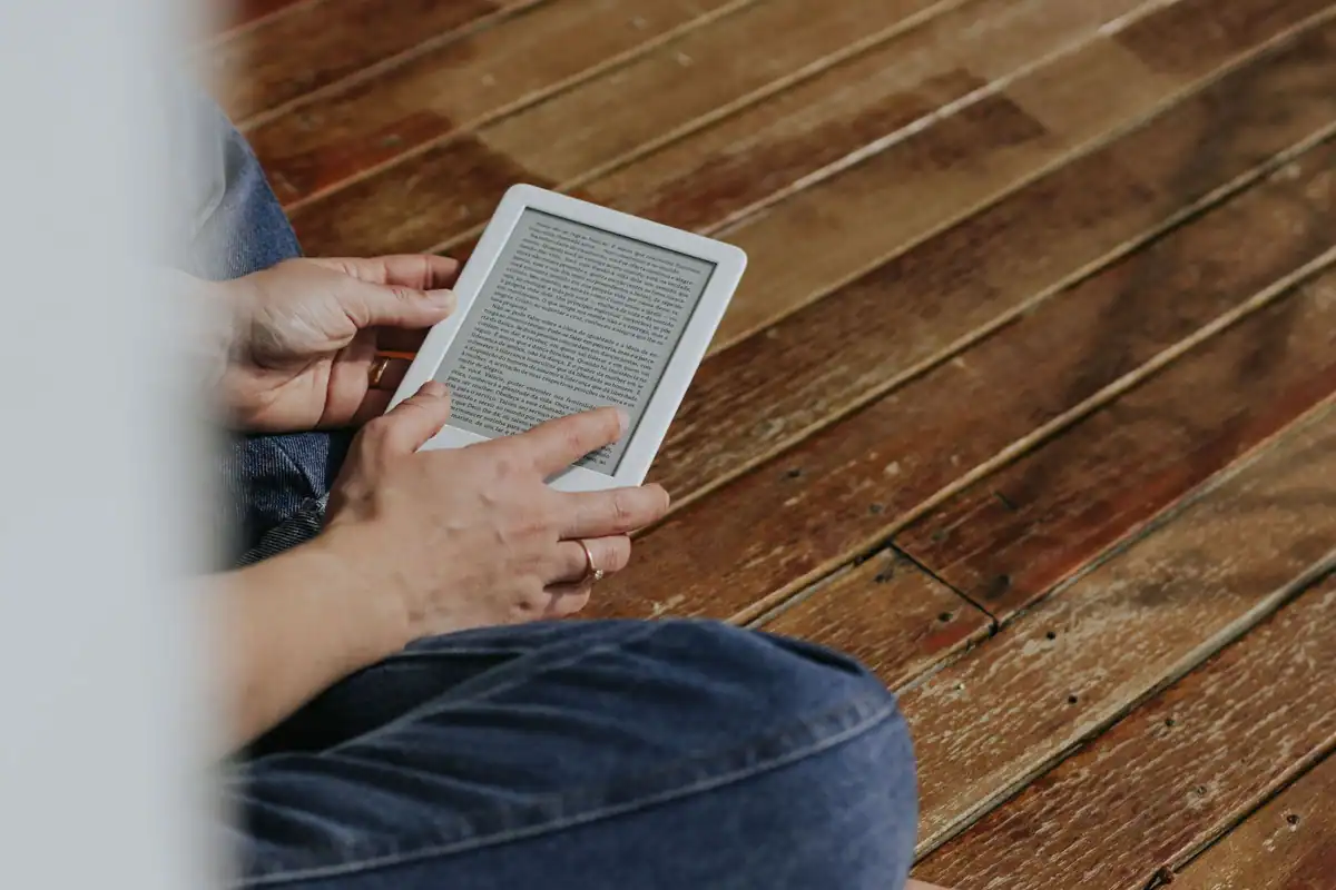 Persoană așezată pe podea, care citește de pe un e-reader alb cu ecran mat