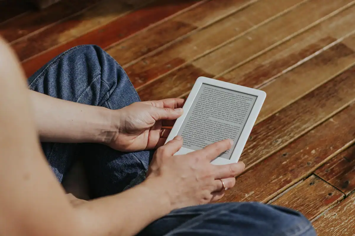 Mâini care țin și ating un e-reader, pe un parchet de lemn închis la culoare