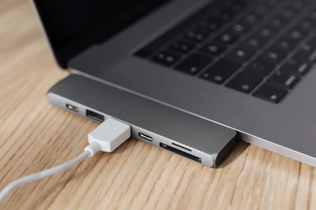 Laptop argintiu pe o masă din lemn, cu un hub USB-C și un cablu alb conectat