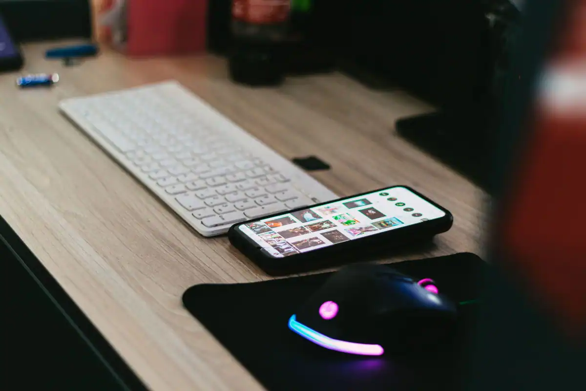 Telefon, tastatură albă și mouse luminat, așezate pe un birou de lemn