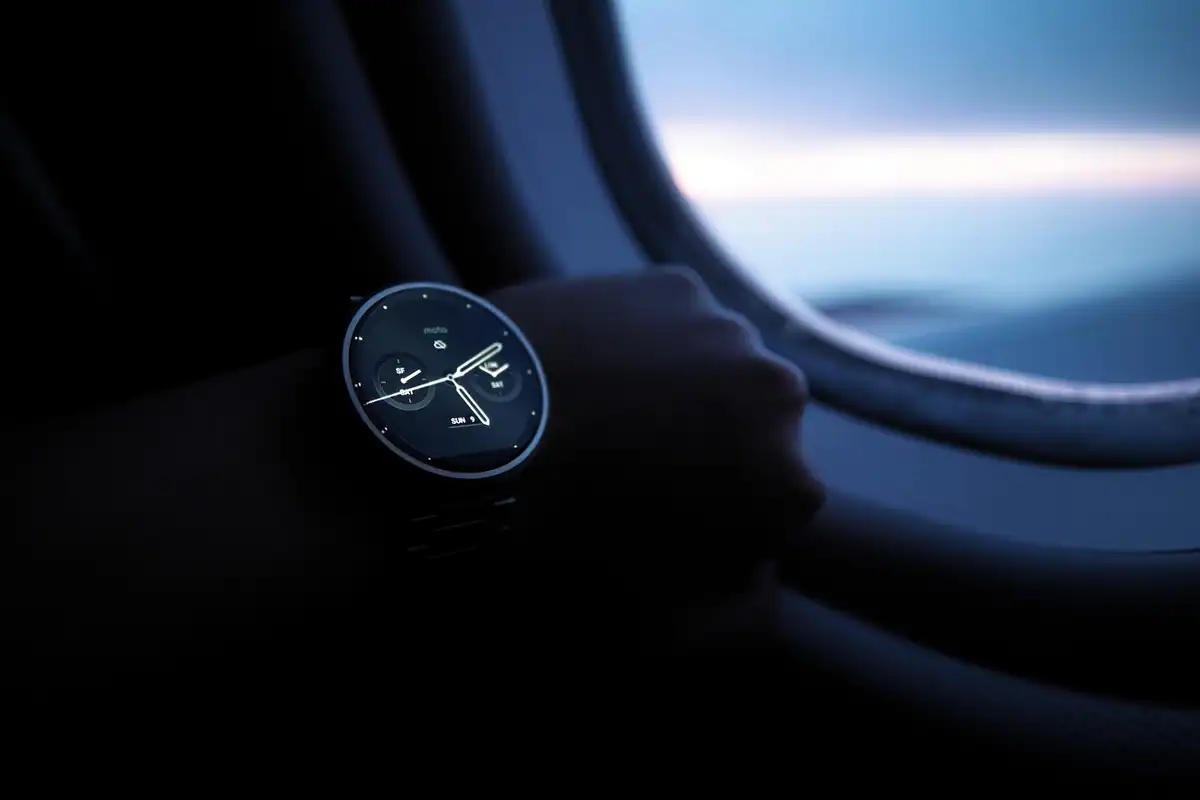 Smartwatch rotund pe mâna unei persoane, lângă geamul întunecat al unui avion