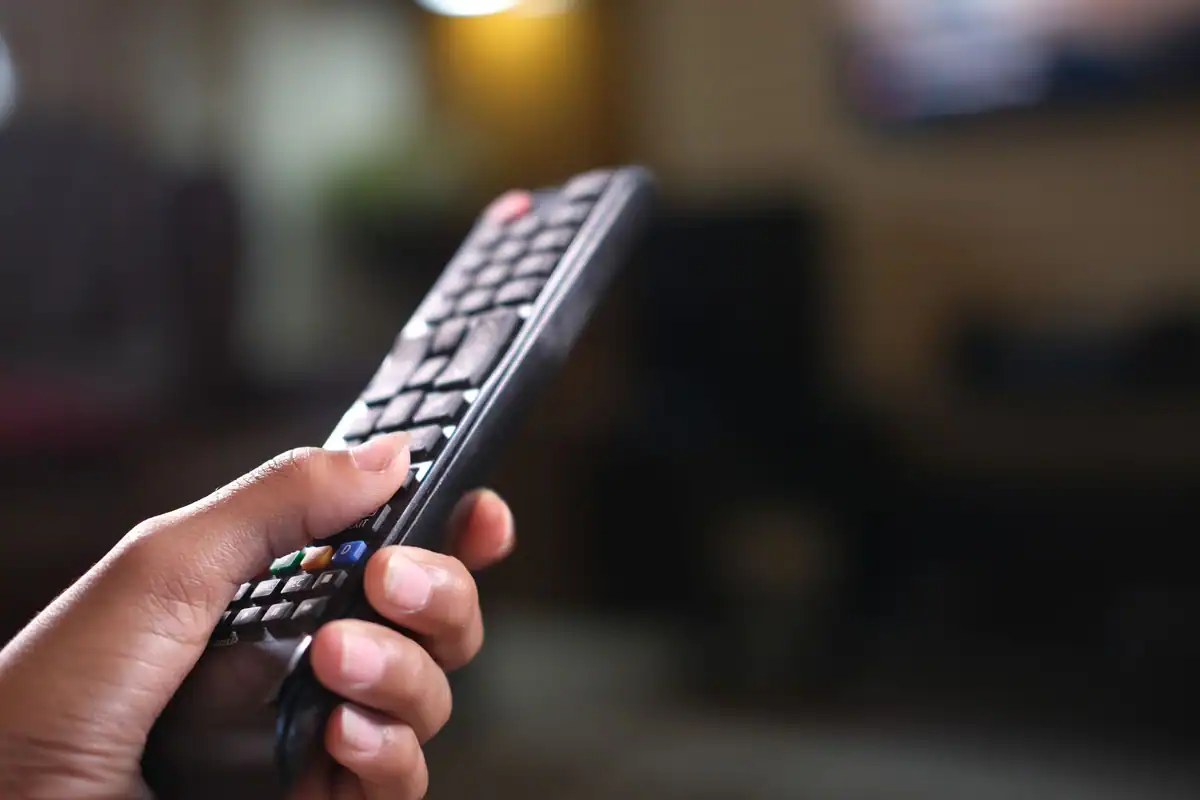 Mână care ține o telecomandă de televizor, într-o cameră slab luminată